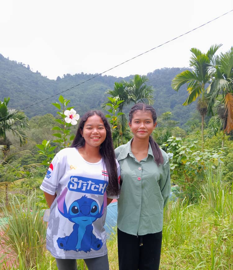 Two young women standing together outdoors with hills and greenery in the background.