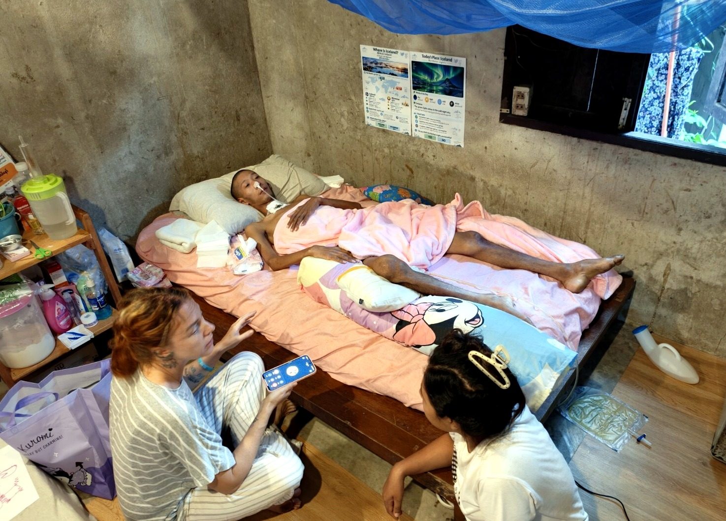 Kin Nage's one-room home: his bed, care supplies on a shelf, and two people sitting beside him.